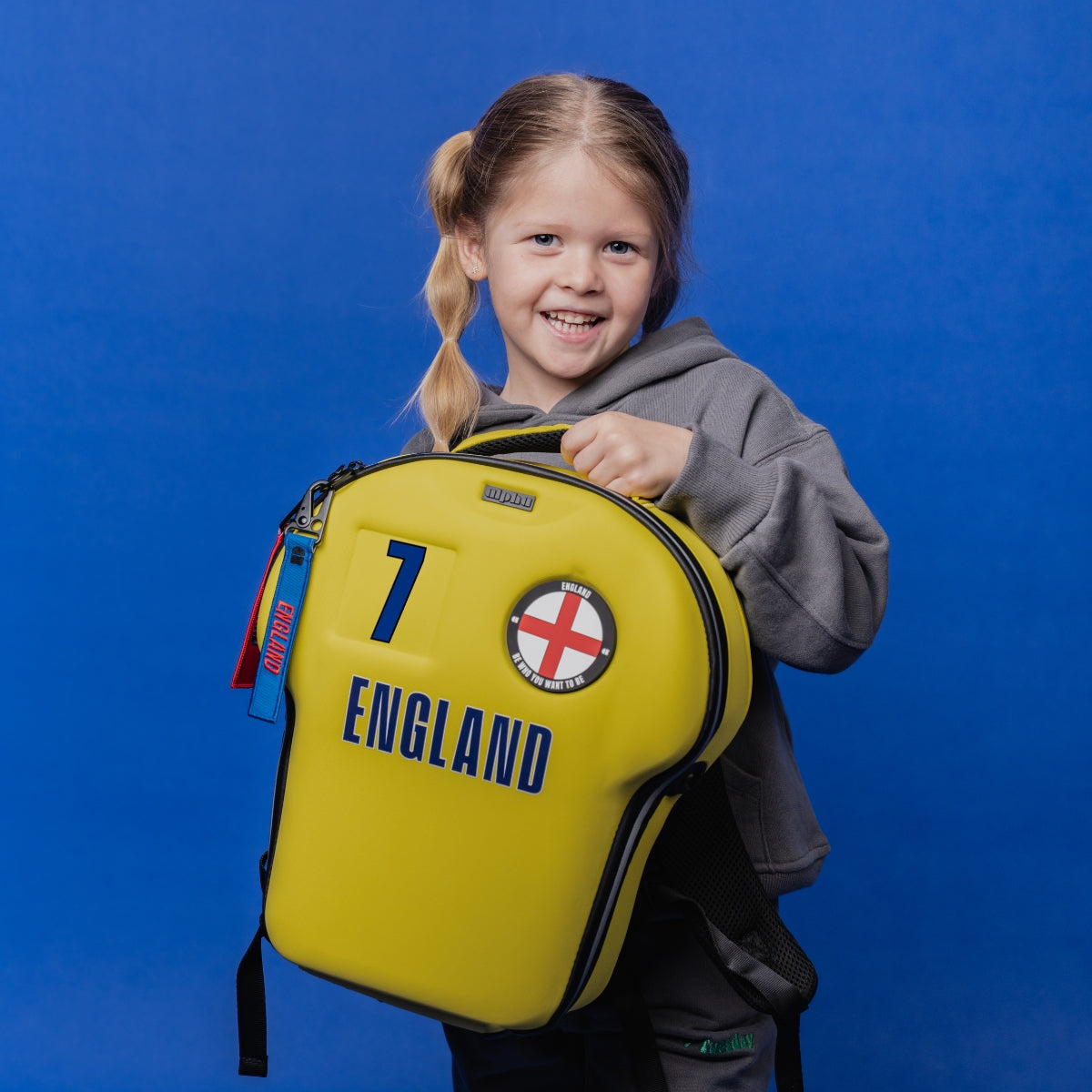 A young girl with blonde pigtails smiling while holding a bright yellow England football-themed backpack against a solid blue studio background.