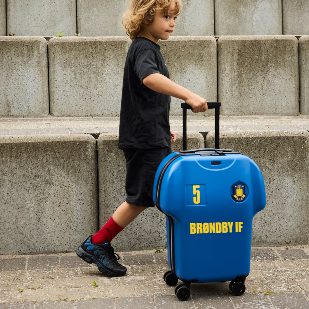 Child pulling a blue suitcase with 'Brondby IF' branding against a concrete wall.