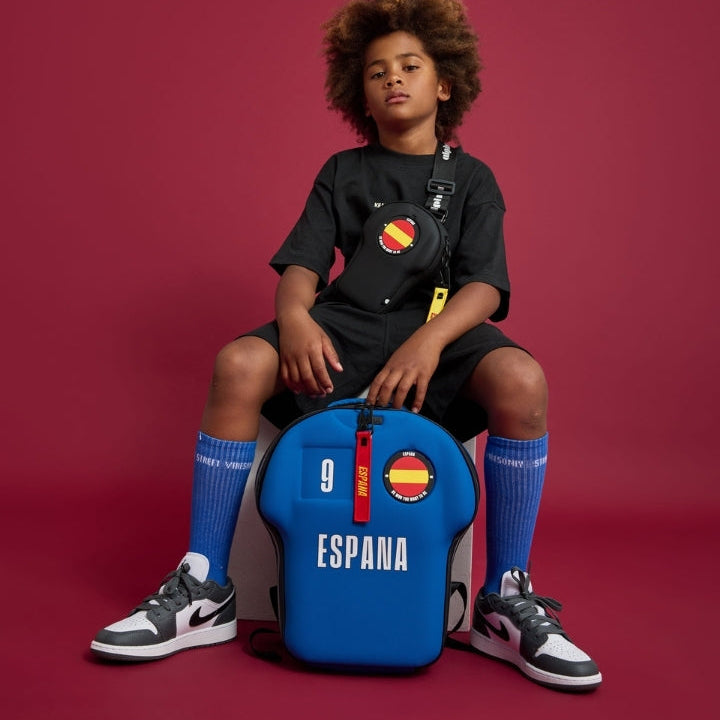 Child sitting on a blue backpack with 'España' branding against a red background