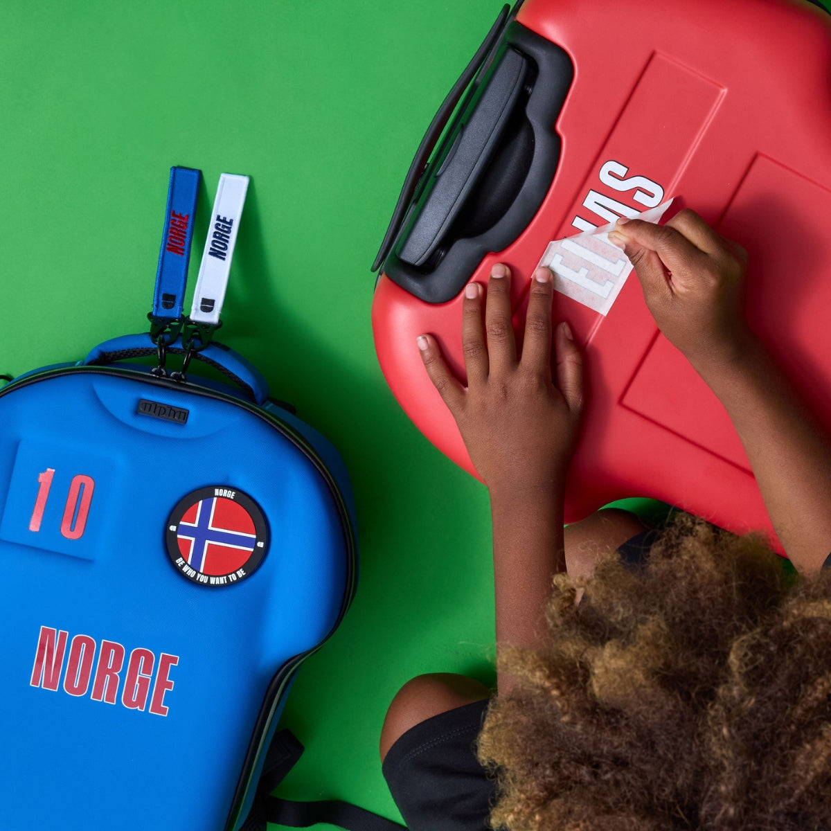 A child's hands are shown peeling a protective sticker off a bright red hard case. In the foreground, a blue hard case with 'NORGE' and a Norwegian flag patch is visible on a solid green background.