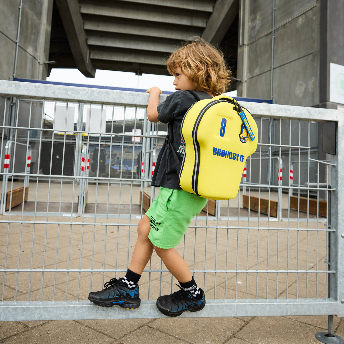 Child with a yellow backpack standing near a metal barrier outdoors.