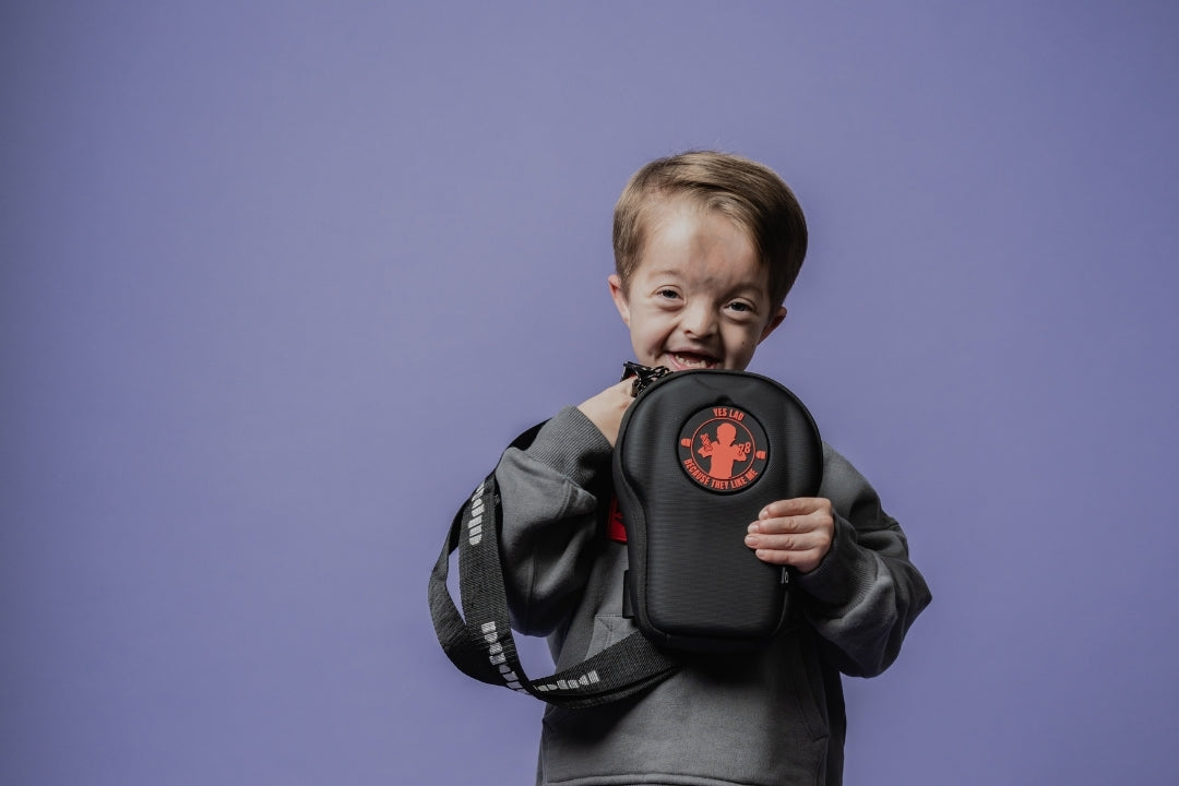 A young boy stands against a lavender studio background holding a black oval-shaped bag with a red cross logo. He is wearing a gray sweatshirt and jogger pants, with a bright blue backpack featuring 'NEDERLAND' and the number 4 placed beside him.